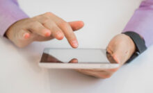 Personne utilisant une tablette tactile avec un doigt posé sur l'écran, portant une chemise violette et une montre noire